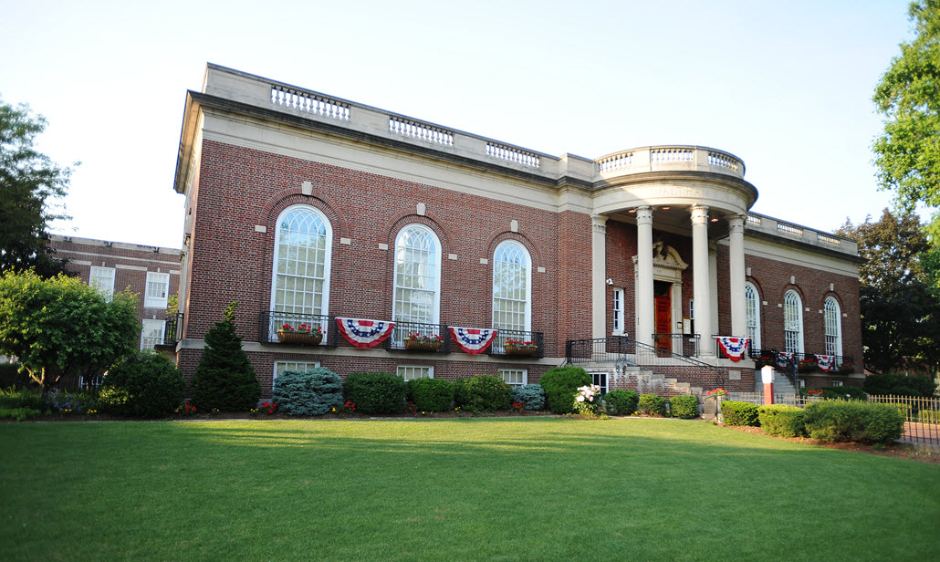 A picture of a large, brick building with columns, surrounded by green grass and bushes. The building is the Waltham Public Library in Waltham, Massachusetts.