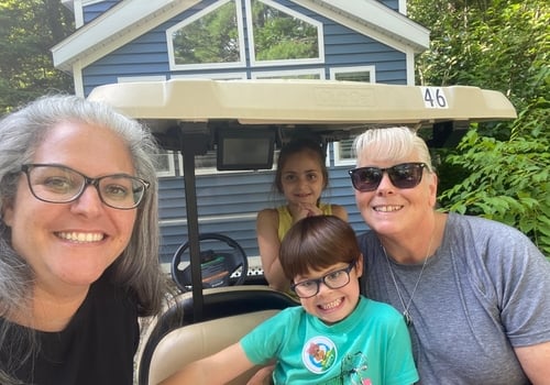 Family of two adults and two kids smiling while sitting on a golfcart in front of their cabin at Point Sebago Resort