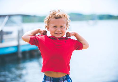 Kid with blonde curly hair and red shirt flexing arm muscles