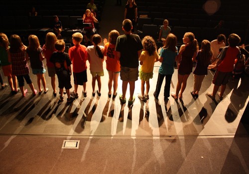 Children standing in a line on a stage, viewed from behind them