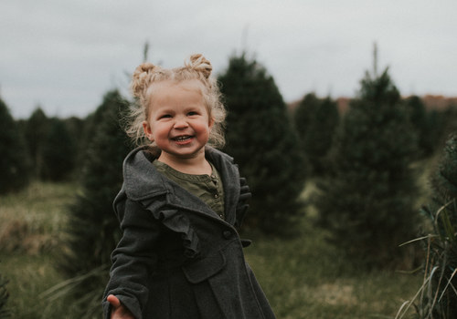 toddler girl looking past the camera with a bright smile and tree farm on cloudy day in background.