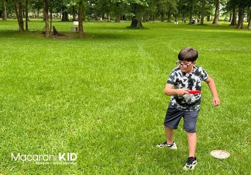 Young boy hold a disc on green grass aiming at a concrete disc golf bucket.
