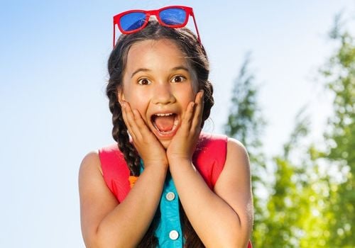 Young girl outside with sunglasses on her head and hands on her face with a surprised & happy expression.