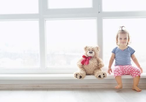 girl and her teddy bear sitting in a large window, which is not covered.