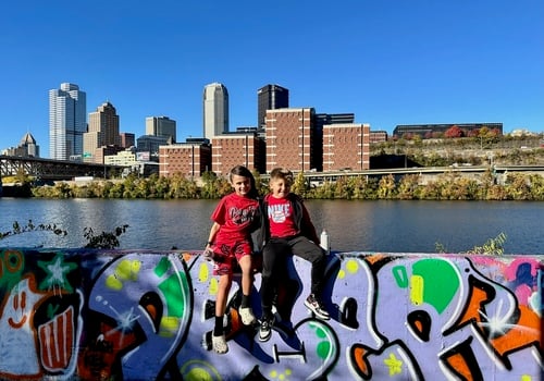 Two boys sitting on a graffiti filled wall with the river and Pittsburgh skyline behind them