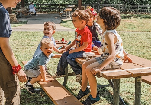 Educator on left side of frame with kids or varying ages on picnic table outdoors