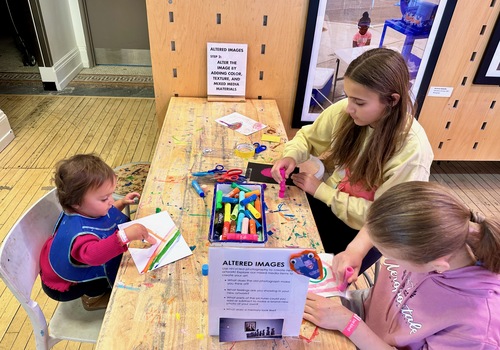 Two school age children and toddler using colorful art supplies at a wooden craft table
