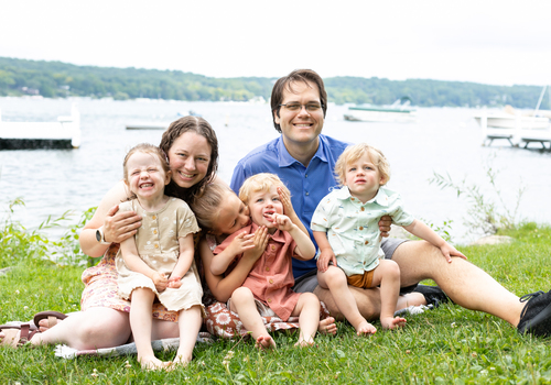 Genevieve and her family of four kids and husband, sitting on grass with a lake in the background