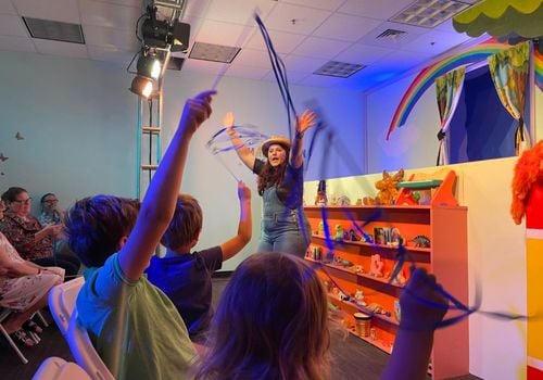 Children wave streamers in the air while watching a performer sing in front of a puppet stage