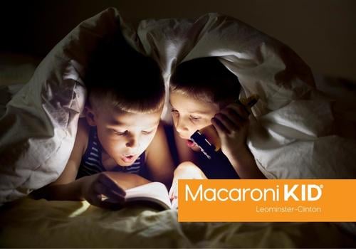 Two young kids reading a book with a flashlight under a blanket.