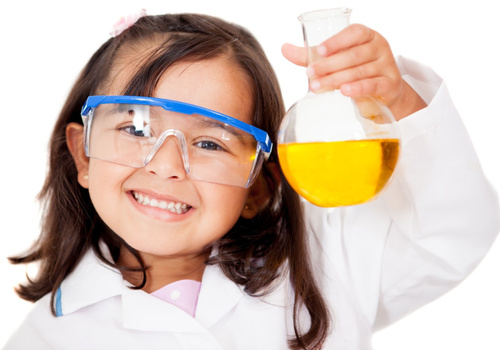 A young girl with dark brown hair is wearing protective science goggles and a lab coat and is holding up a science beaker containing a yellow liquid. She is smiling.