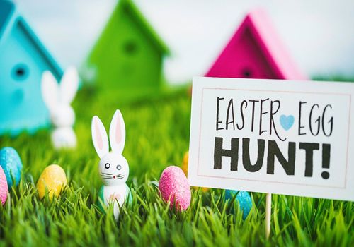 Shows easter eggs and a wooden bunny in the grass with a sign that says easter egg hunt