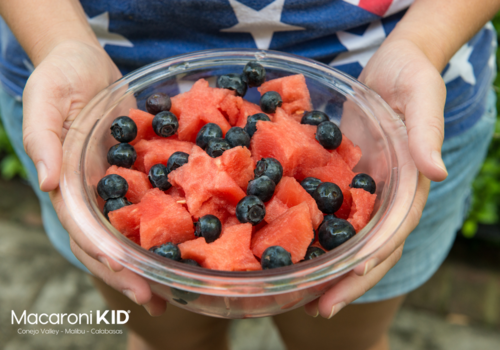 person wearing a patriotic shirt holding out a glass bowl with star shaped watermelon and blueberries