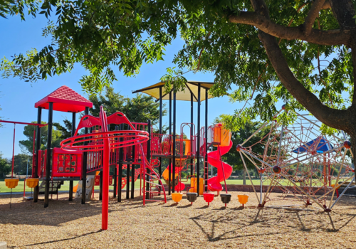 Tree-framed view of various red and yellow themed playground structures at DeGarmo Park