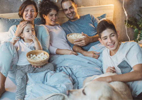 A happy Caucasian family enjoys a movie night on their bed, surrounded by love, popcorn, and their adorable Yellow Labrador.