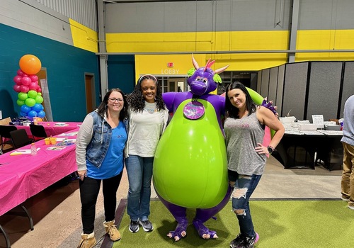 Three women, Macaroni KID publishers stand up to pose with a large, purple, inflatable dragon with a green belly