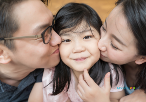 man and woman (presumably parents) kissing little girl on each cheek. Girl grinning and looking at camera.