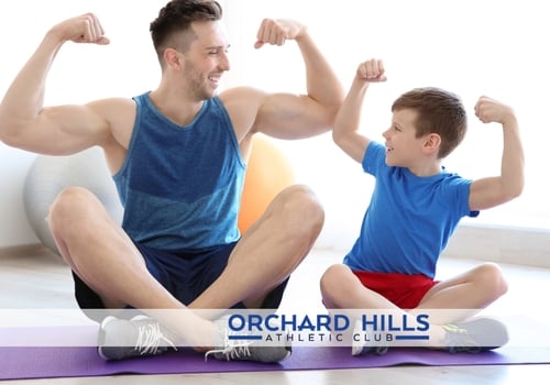 Man and boy smiling and flexing their muscles while sitting on a yoga mat.