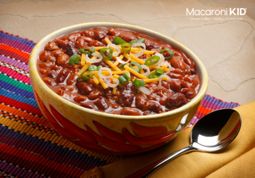 A bowl of chili topped with cheese and and green onions on a table with a bright colored striped placement and a spoon