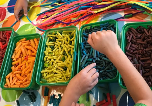 Green plastic trays each with a different color macaroni. Childs hand reaching to make a necklace with noodles and a pipe cleaner.