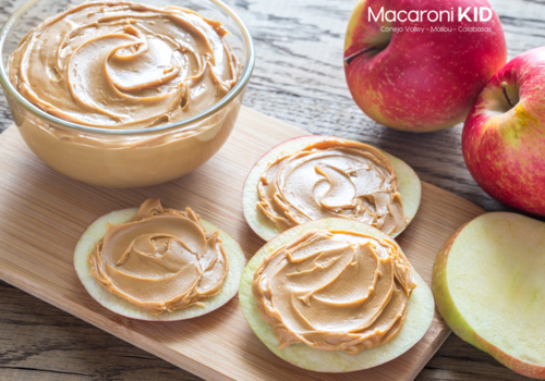 Apple slices with peanut butter on a wood board with whole apples and a bowl of peanut butter