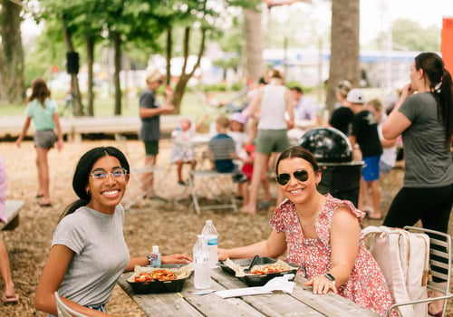 two ladies smiling at the camera eating lunch at a picnic table