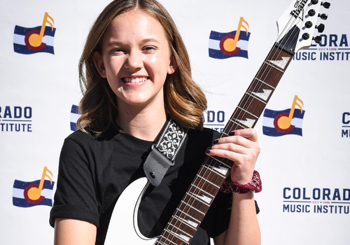 student holding guitar and posing in front of Colorado Music Institute banner