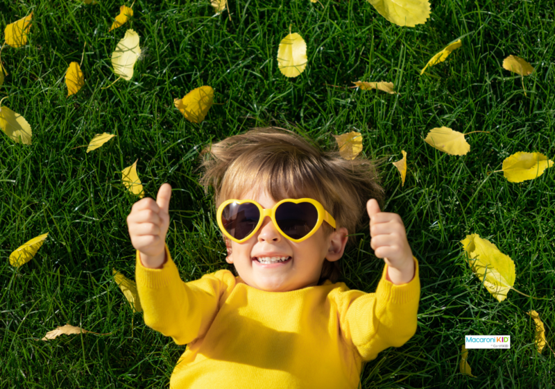 Kid lying on green grass giving thumbs up. wearing bright yellow.