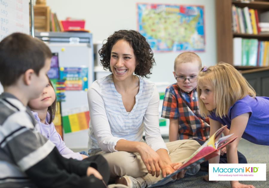 A young teacher with tan skin and curly hair reads to a group of caucasian children.