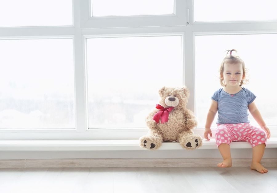 girl and her teddy bear sitting in a large window, which is not covered.