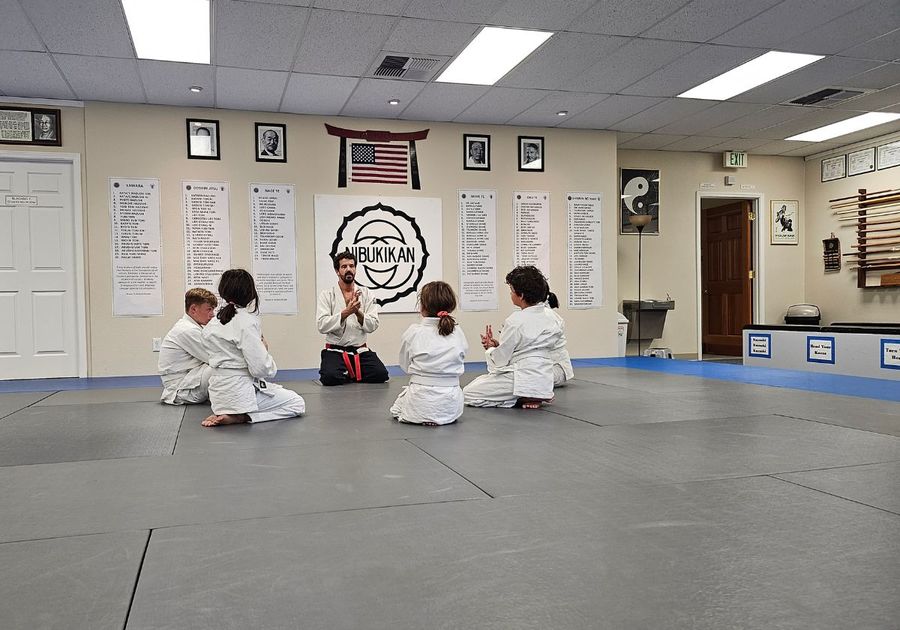 5 Kids kneel in a circle facing their instructor, stretching their wrists in a jujitsu class
