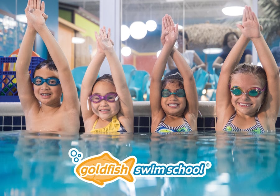 4 children hold arms above head in dive position while wading in a pool.  Smiles and googles on all of them.