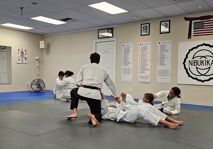 5 kids in various positions grappling, laughing, and learning jujitsu from their instructor who is kneeling and facing away from the camera