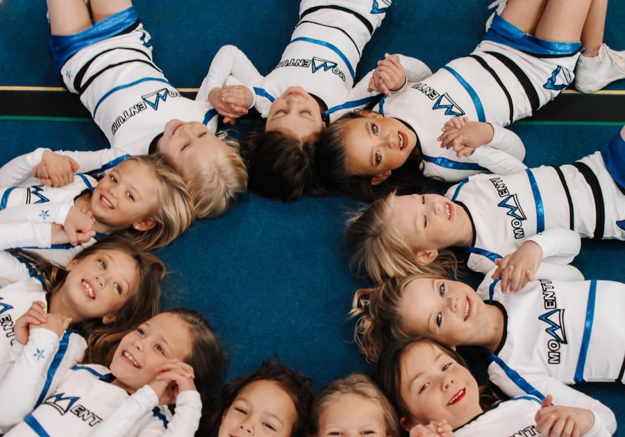 11 young cheerleaders in uniforms that say Momentum. They are lying on the ground with their heads together to form a circle.