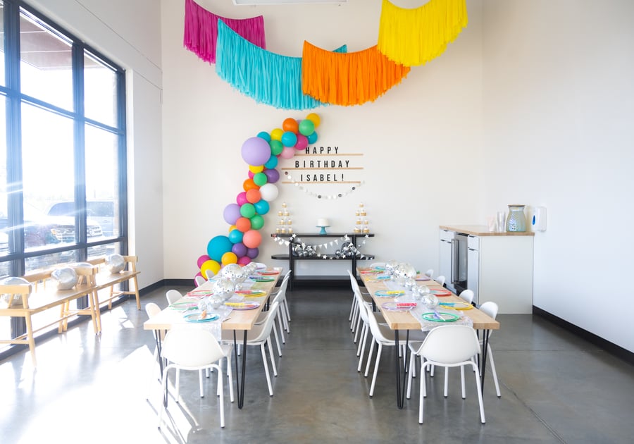 Birthday party room with bright sunlight from floor to ceiling window with two rectangle tables and brightly colored balloons and banners