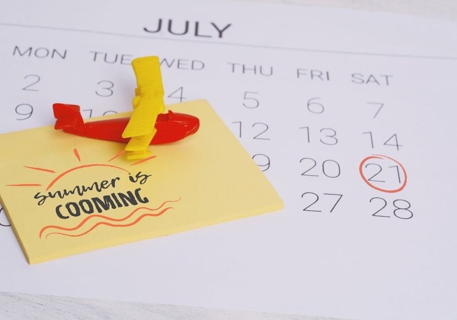 Close up of a July calendar with the date Saturday 21st circled in red. There are yellow Post-it notes that say summer is coming stacked on the calendar with a red and yellow airplane paperweight.