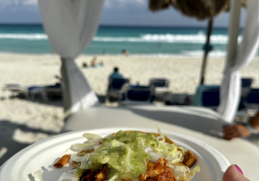 An image of a tropical beach in Cancun, Mexico featuring a turquoise blue sea and someone holding a plate of tacos