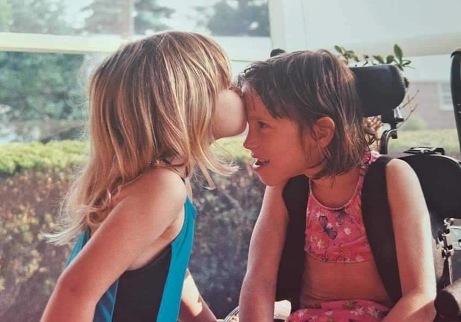 Little girl with brown har in wheelchair leaning over to be kissed on the forehead by her sister who is also little.