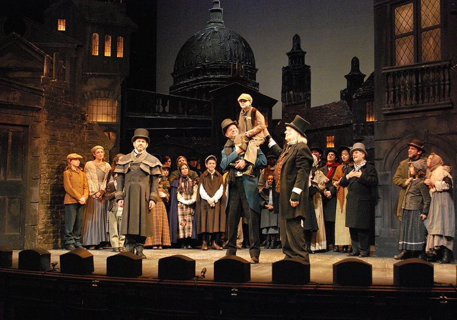 tiny tim on the top of Bob Cratchit's shoulder with Ebeneezer Scrooge in crowd