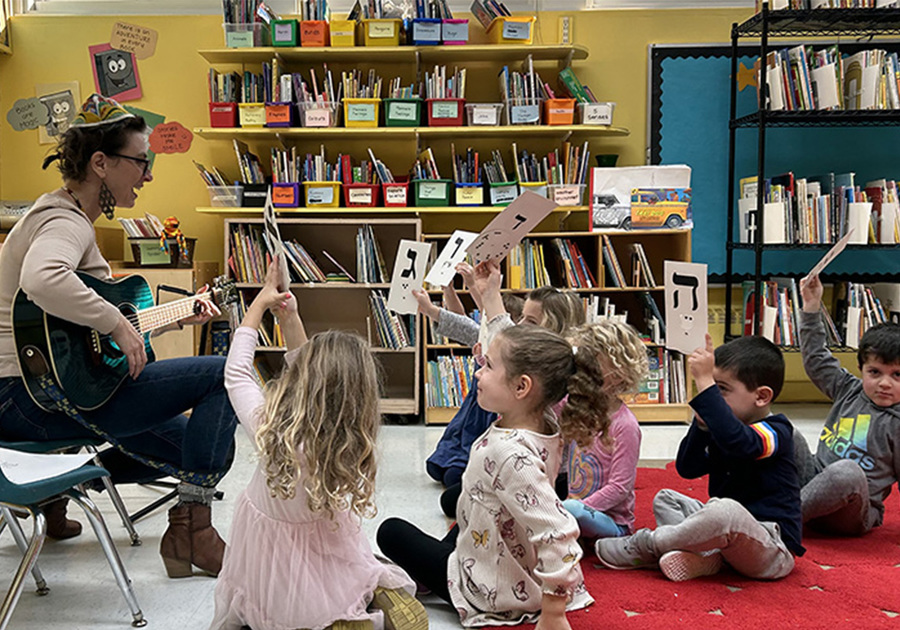 Teacher playing a guitar and singing with kids interacting and raising symbols.