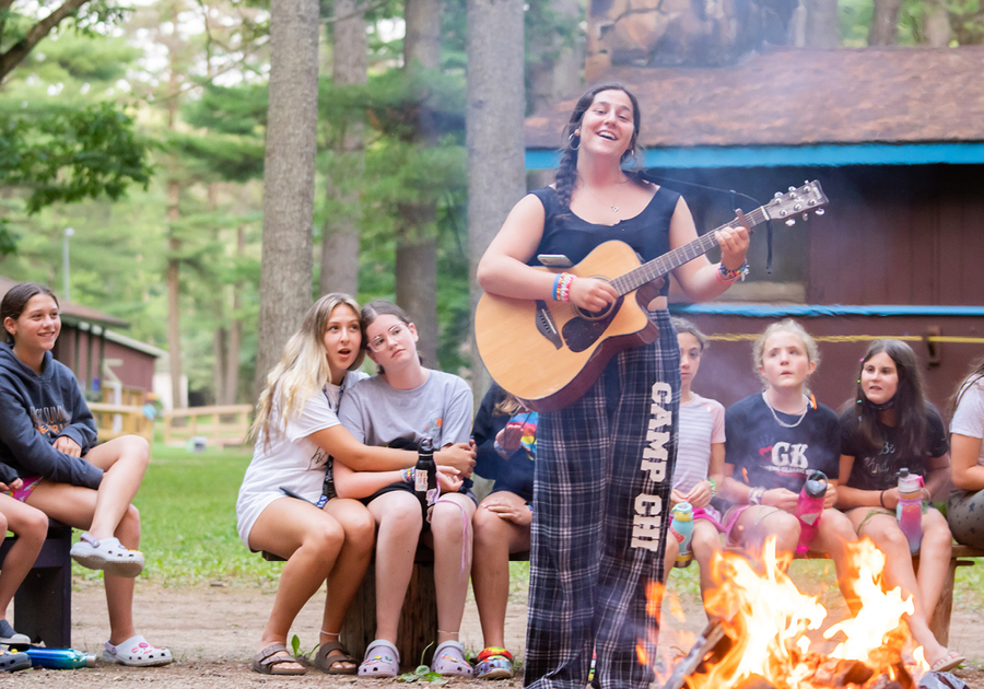 girl playing guitar and singing in front of campers at camp fire
