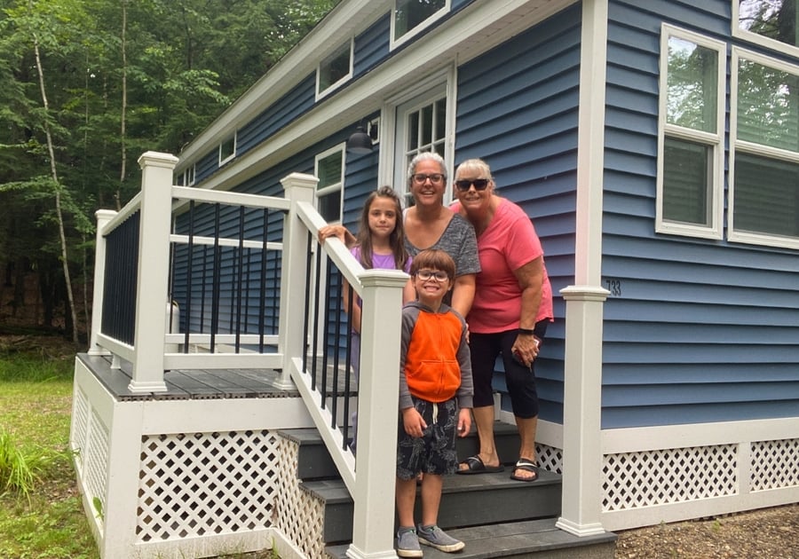 Family with two adults and two kids stand on the front porch of a small cabin at Point Sebago Resort
